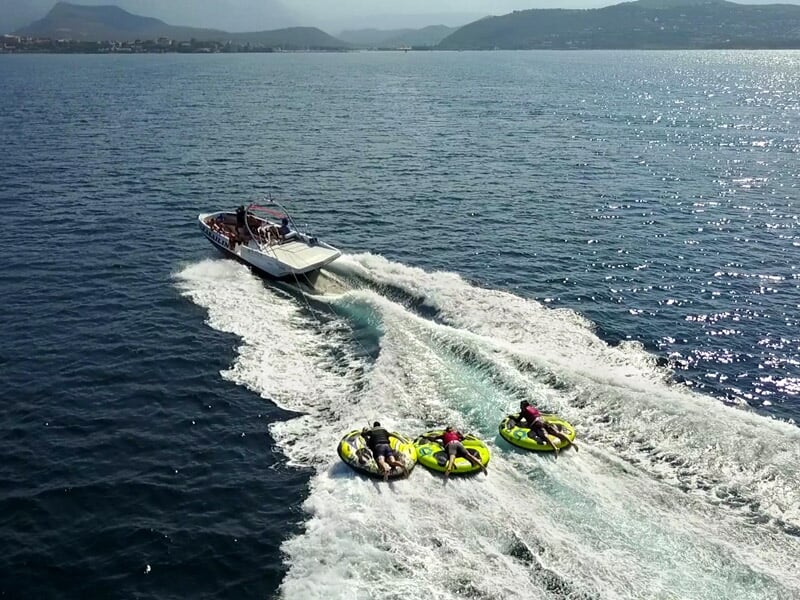 Vue aérienne d'un bateau tirant trois bouées tractées avec des passagers sur une mer calme et montagnes au loin.