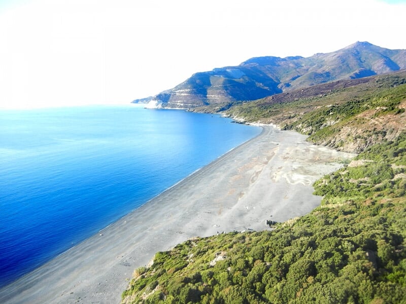 Plage de sable fin bordée par une mer bleue et une végétation dense avec des montagnes en arrière-plan sous un ciel clair.