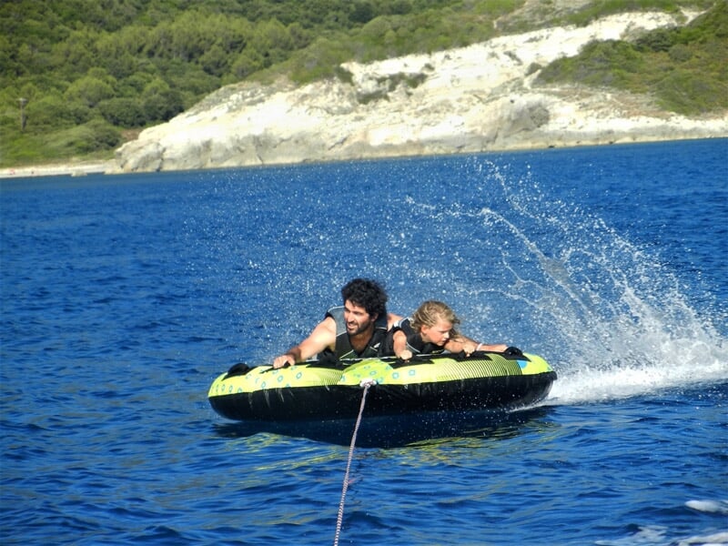 Deux personnes souriantes sur une bouée tractée sur une mer calme, éclaboussures d'eau autour et paysage naturel en arrière-plan.