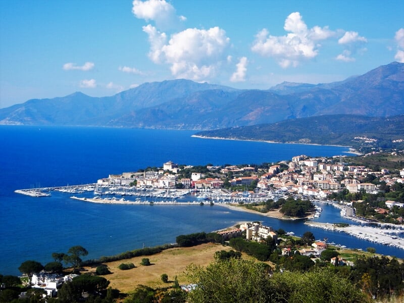 Vue panoramique d'une ville côtière avec un port de plaisance, montagnes en arrière-plan et ciel bleu clair.