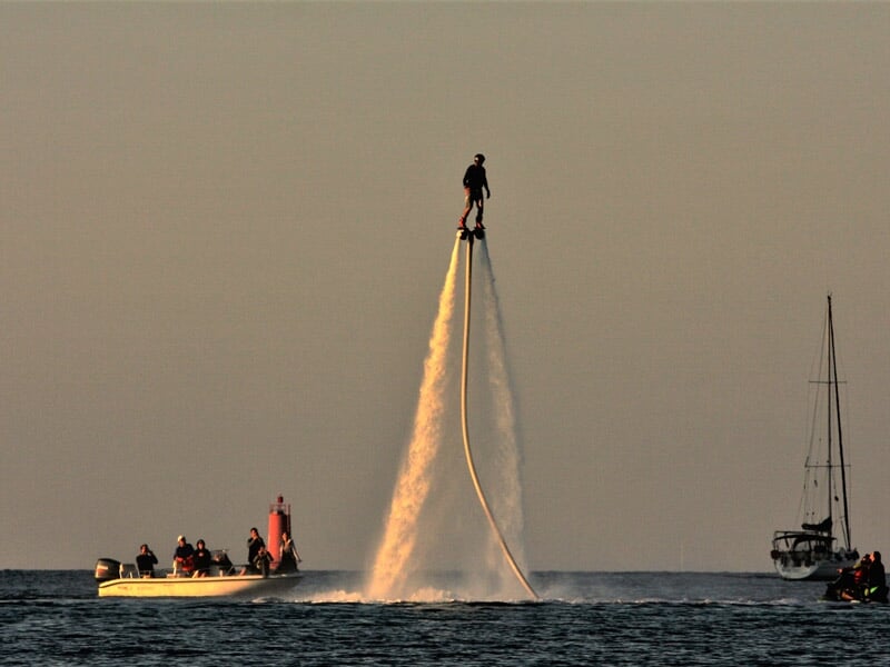 Personne en flyboard s'élevant au-dessus de l'eau, propulsée par un jet d'eau, avec un bateau et d'autres personnes en arrière-plan.