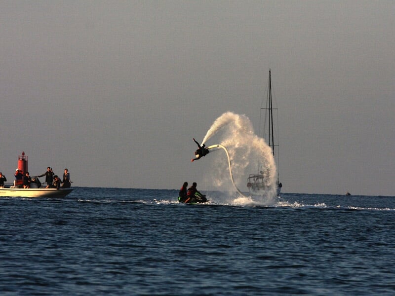 Personne effectuant une figure acrobatique en flyboard au-dessus de la mer, entourée d'eau et de bateaux au loin.