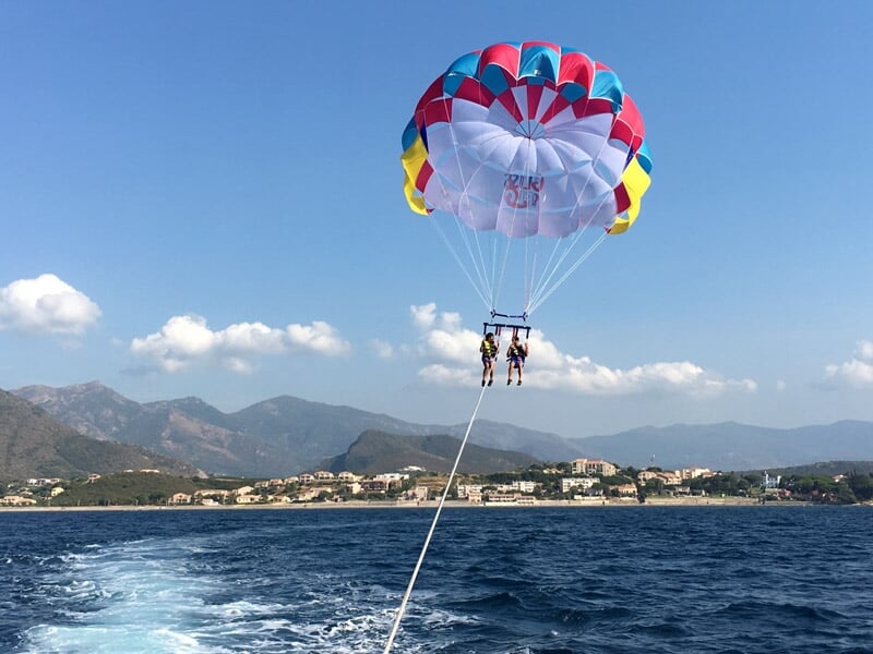 Deux personnes en parachute ascensionnel attachées à un bateau naviguant sur une mer bleue avec montagnes en arrière-plan.