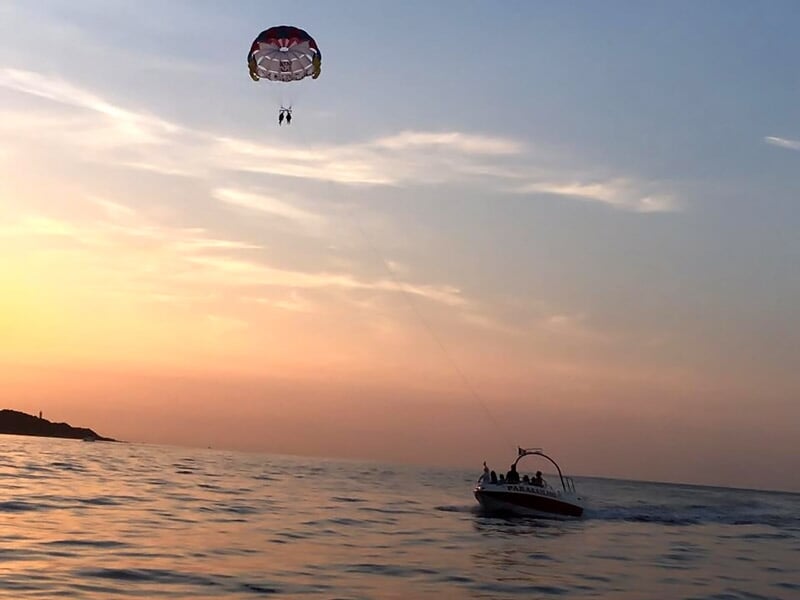 Parachute ascensionnel coloré avec deux passagers au-dessus de la mer, bateau tirant le parachute visible au loin.
