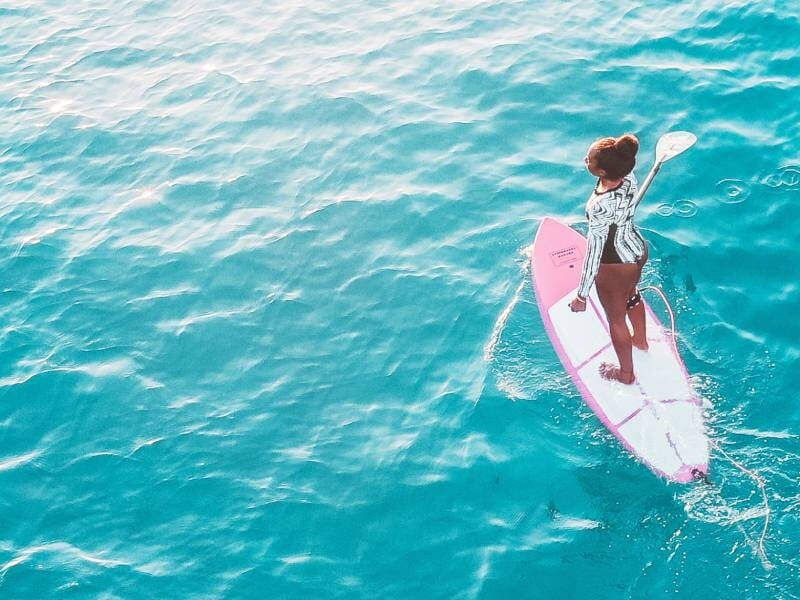 Femme debout sur un stand-up paddle rose pagayant sur une eau turquoise claire sous un ciel ensoleillé.