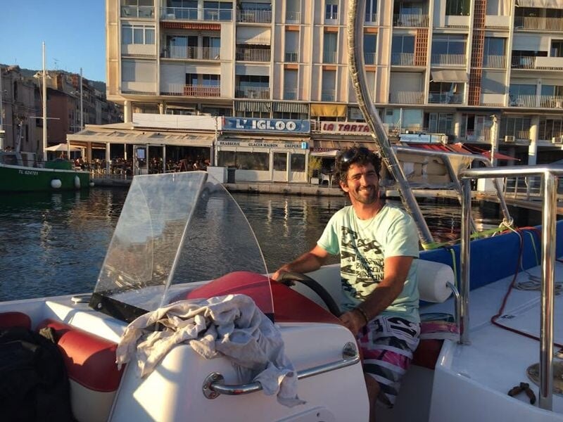 Un homme souriant à bord d'un bateau amarré dans un port avec des bâtiments en arrière-plan au coucher du soleil.