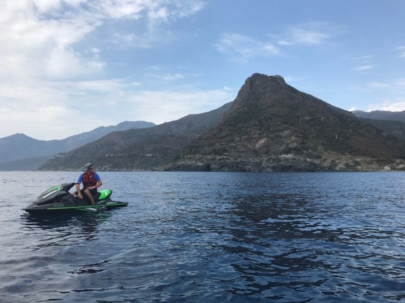 Homme en gilet de sauvetage orange et casque, assis sur un jet ski noir et vert, avec une grande montagne rocheuse et la mer bleue en arrière-plan sous un ciel partiellement couvert