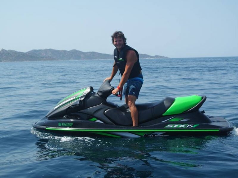 Homme souriant, debout sur un jet ski noir et vert, portant un gilet de sauvetage noir et des shorts rayés, avec des montagnes lointaines et une mer calme