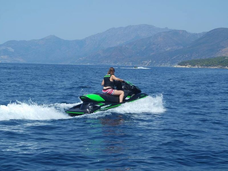 Personne en maillot de bain à rayures rouges et blanches conduisant un jet ski noir et vert sur une mer bleue calme avec des montagnes en arrière-plan