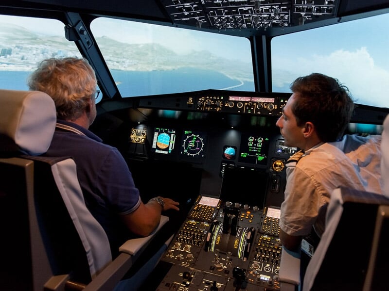 Deux hommes dans un simulateur de pilotage d'avion, discutant et regardant les écrans de contrôle et la vue extérieure.