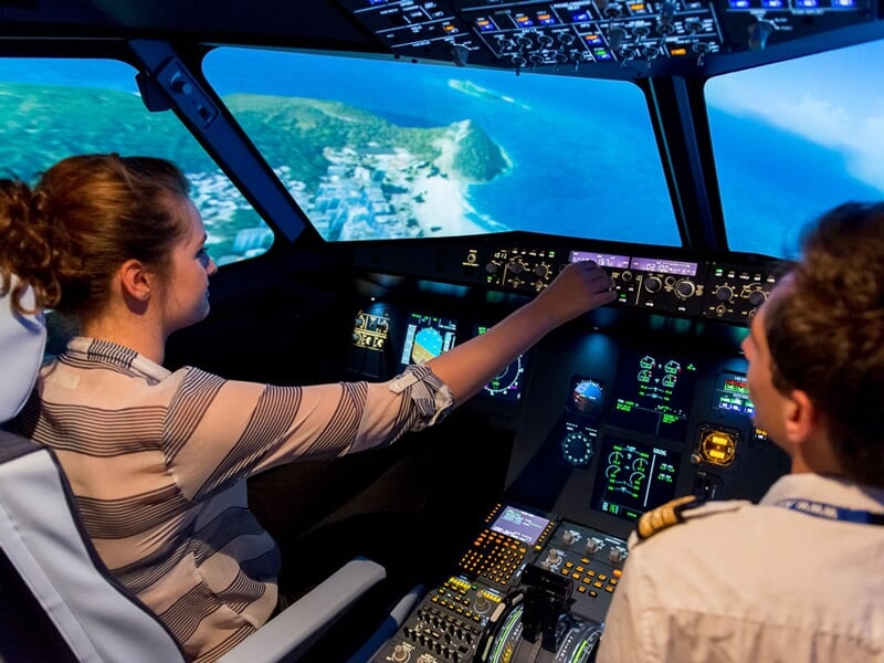Femme et homme dans un simulateur d'avion, la femme manipule les commandes avec un paysage côtier visible à l'extérieur.
