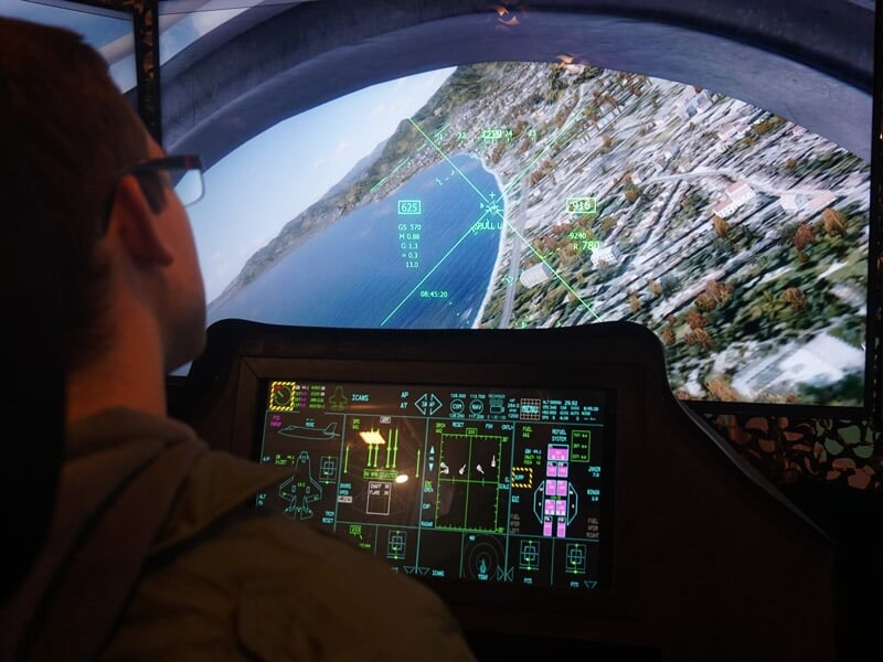 Homme dans un simulateur de vol militaire, regardant un écran avec une vue aérienne et des données de vol.