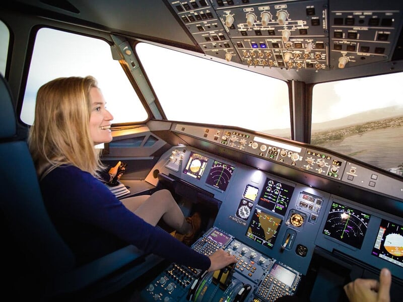 Femme souriante aux commandes d'un simulateur de vol, avec un paysage montagneux visible à travers le cockpit.