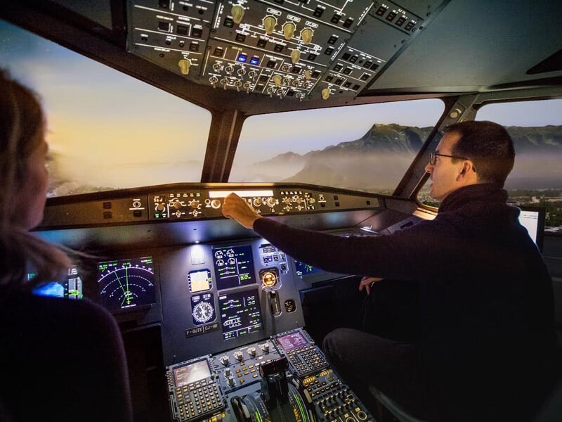 Homme et femme dans un simulateur d'avion, observant les instruments et le paysage montagneux au coucher du soleil.