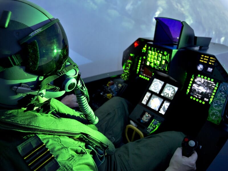 Cockpit de simulateur de vol F-35 avec pilote portant un casque et des lunettes de vision nocturne, panneaux de contrôle lumineux en vert