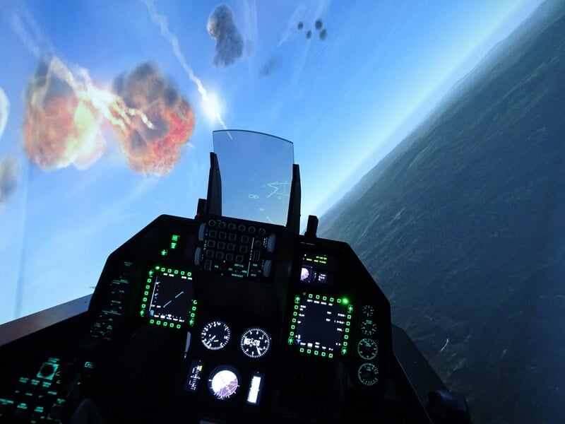 Vue depuis un cockpit de simulateur de vol de chasse, avec explosions visibles à l'extérieur et instruments lumineux.
