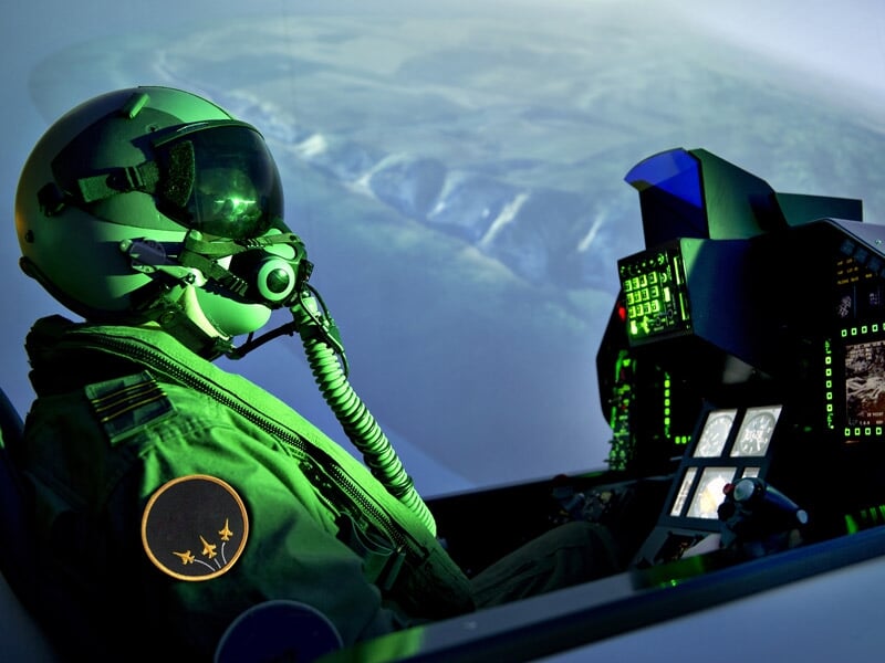 Simulateur de vol F-35 montrant le cockpit illuminé par des écrans verts, avec un pilote en combinaison de vol et casque, vue sur des montagnes depuis le cockpit
