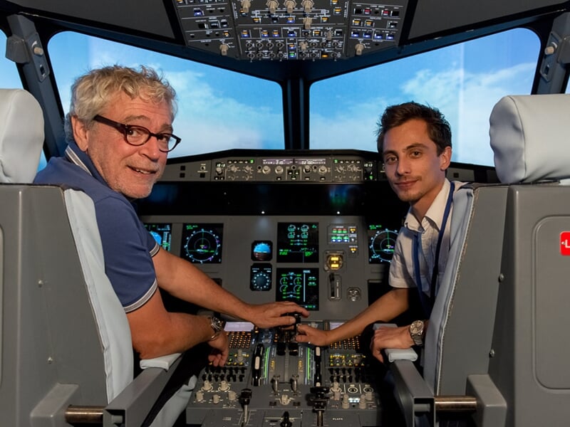 Deux hommes dans un simulateur de vol, souriants et manipulant les commandes dans un cockpit réaliste.