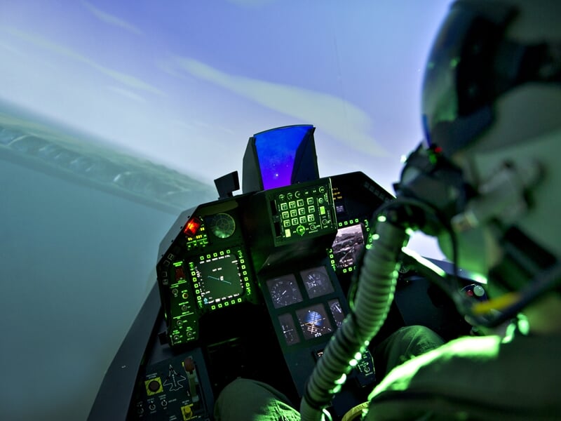 Simulateur de vol F-35 avec vue de l'intérieur du cockpit, les écrans affichant les données de vol et le pilote en action, le ciel à l'extérieur