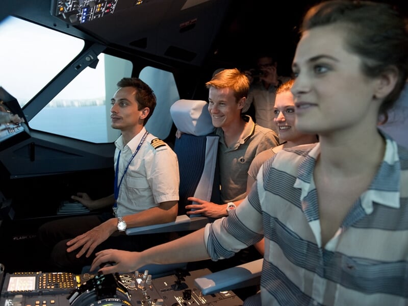 Groupe de jeunes dans un simulateur d'avion, une femme aux commandes et plusieurs personnes regardant autour.