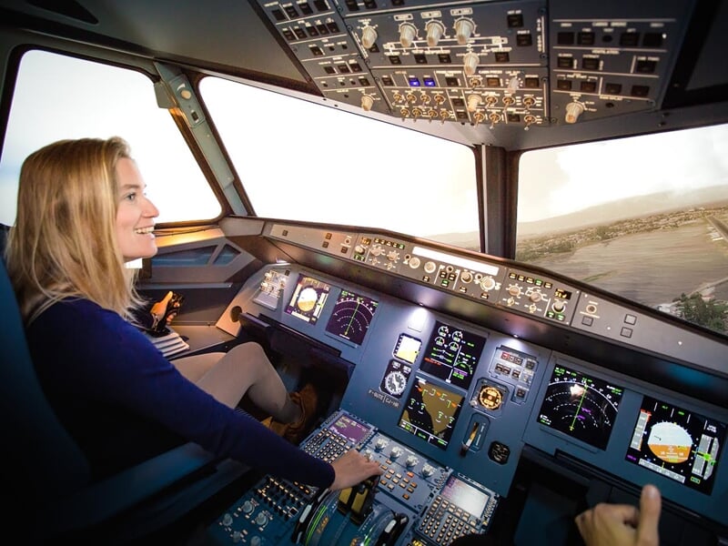 Femme souriante aux commandes d'un simulateur de vol, paysage urbain visible à travers le pare-brise du cockpit.