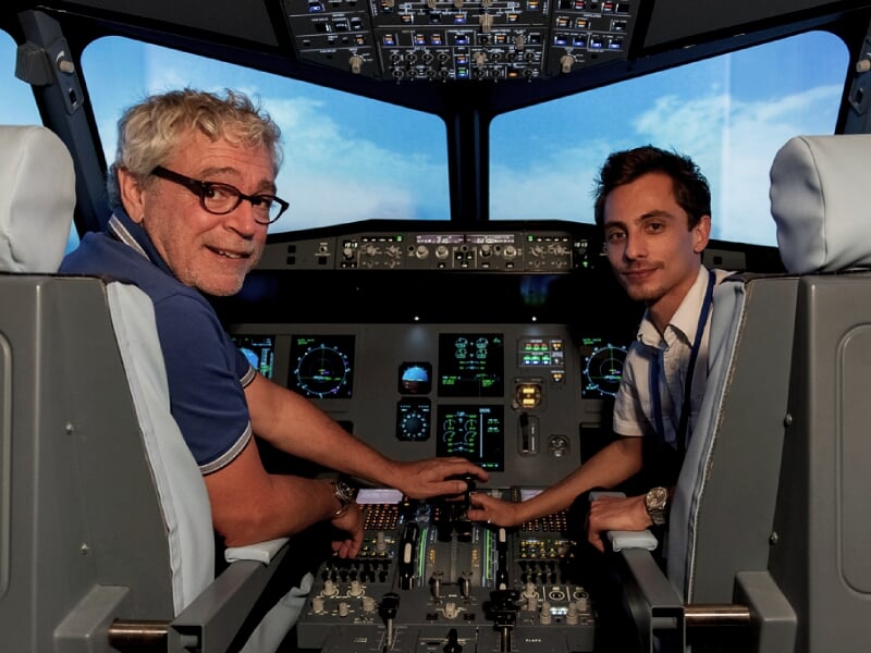 Deux hommes souriants, un jeune et un plus âgé, assis dans un simulateur de cockpit d'avion de ligne, manipulant les commandes