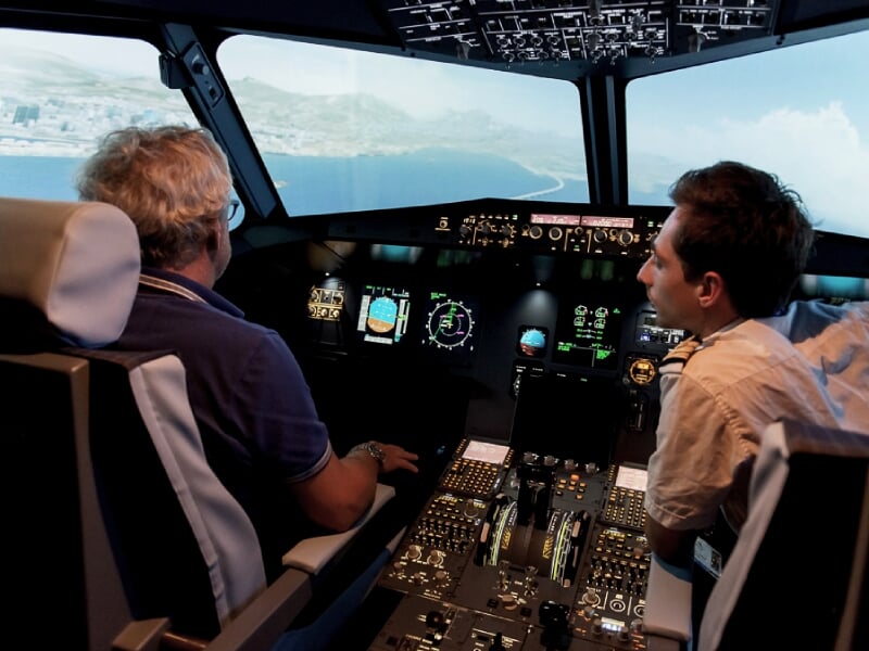 Deux hommes dans un simulateur de vol, observant les instruments et la vue extérieure d'un paysage côtier.