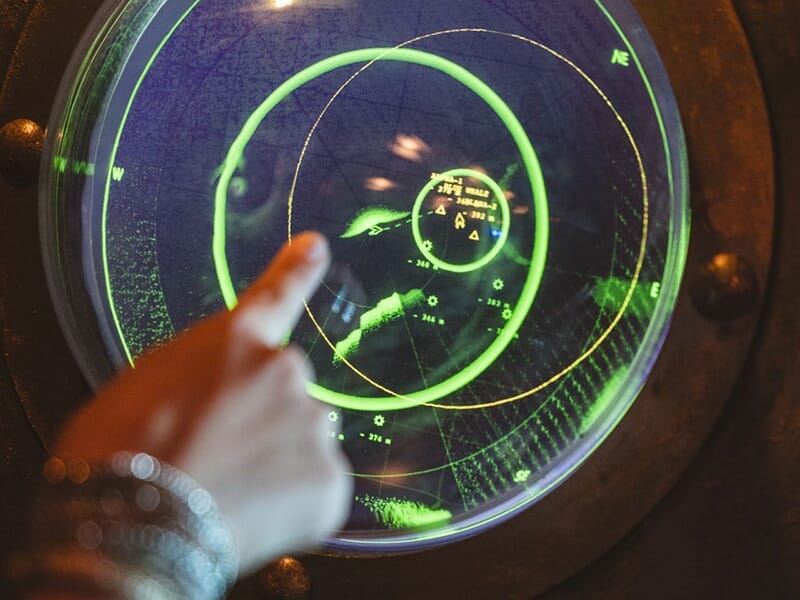 Doigt pointant vers un écran radar circulaire affichant des informations lumineuses en vert.