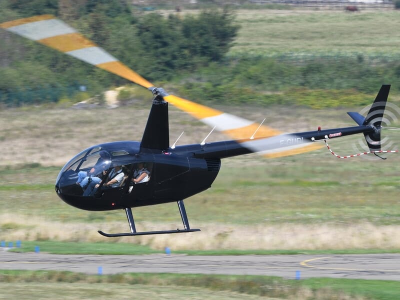 Hélicoptère noir en vol au-dessus d'une piste d'aéroport avec un pilote visible à l'intérieur
