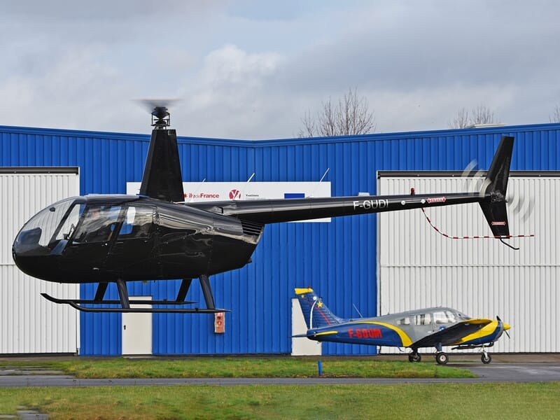 Hélicoptère noir au sol devant un bâtiment bleu avec un petit avion visible en arrière-plan