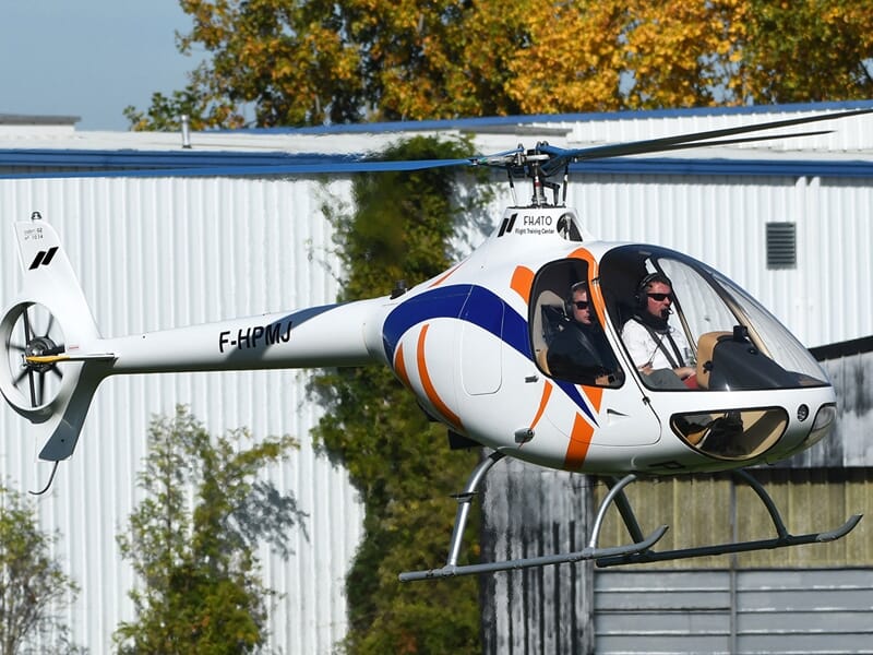 Hélicoptère blanc avec motifs bleus et orange en vol près d'un bâtiment, pilote visible à bord à Toussus-le-Noble.
