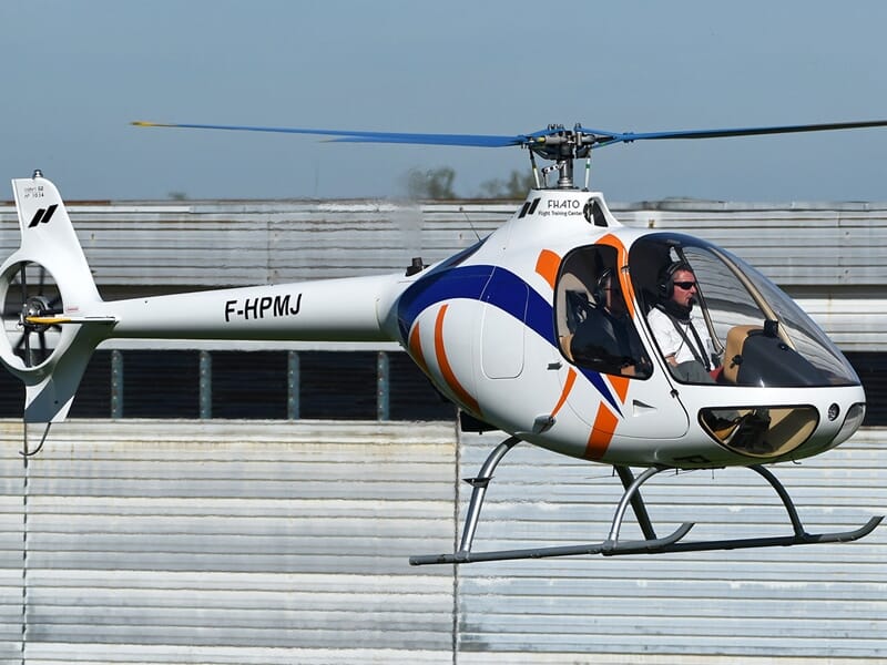 Hélicoptère blanc avec motifs bleus et orange en vol stationnaire devant un hangar, pilote portant des lunettes de soleil.
