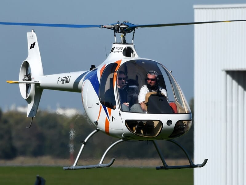 Hélicoptère blanc avec motifs bleus et orange en vol près d'un bâtiment blanc, deux personnes à bord avec lunettes de soleil.