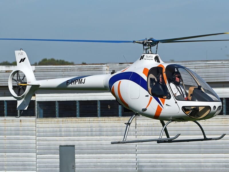 Hélicoptère blanc avec motifs bleus et orange en vol stationnaire devant un hangar métallique, pilote visible à bord.