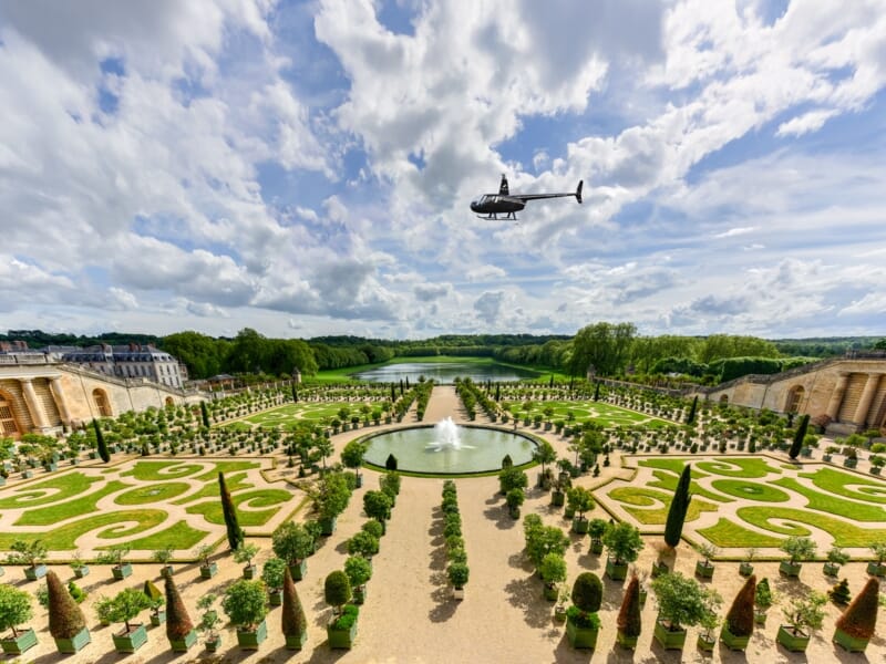 Vue aérienne d'un hélicoptère survolant un jardin à la française avec fontaine et allées symétriques sous un ciel nuageux.