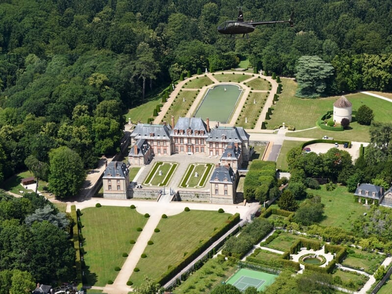 Hélicoptère volant au-dessus d'un château entouré de jardins, pelouses et arbres dans un paysage verdoyant et ensoleillé.