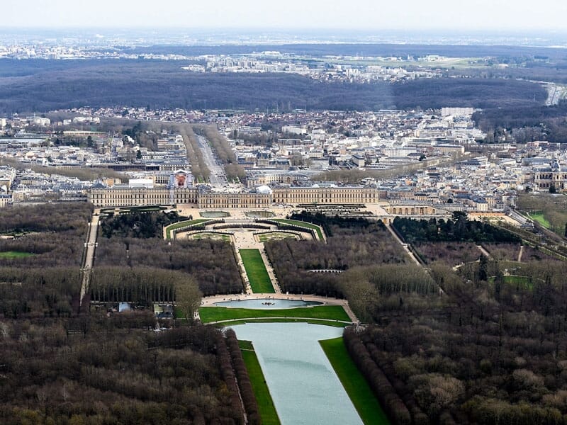 Vue aérienne du château de Versailles avec ses jardins et bassins, entouré de la forêt et de la ville au loin.