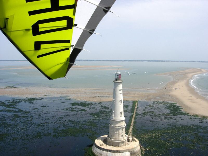 Vue aérienne d'un phare blanc sur un îlot entouré d'eau avec une aile d'ULM jaune visible en haut à gauche.