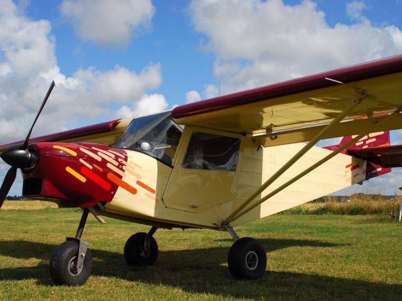 Petit avion ultraléger jaune et rouge stationné sur un champ vert sous un ciel bleu avec quelques nuages blancs.