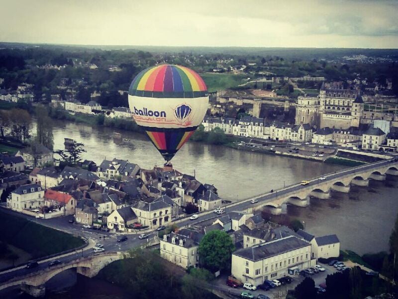 Montgolfière aux couleurs vives flottant au-dessus d'une ville pittoresque traversée par une rivière, avec un pont et des bâtiments historiques visibles en contrebas