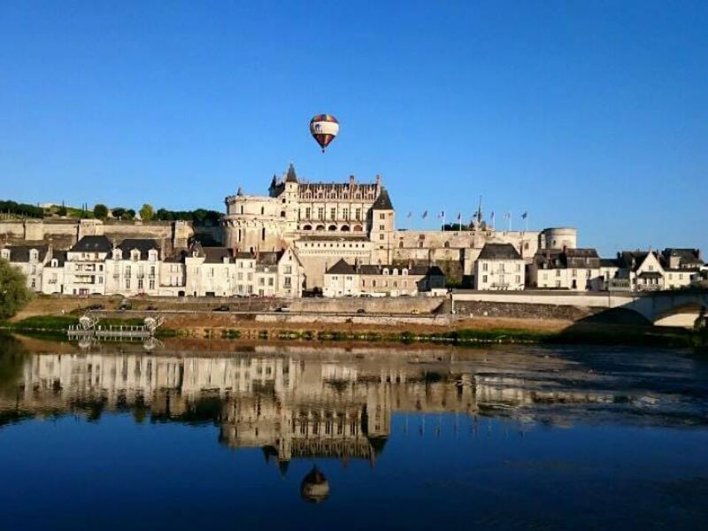 Montgolfière multicolore flottant au-dessus du Château d'Amboise, avec son reflet visible dans les eaux calmes de la Loire, sous un ciel bleu clair