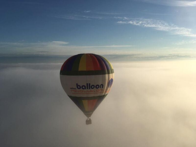 Montgolfière multicolore naviguant en altitude au-dessus d'une mer de nuages, baignée dans une lumière douce, avec un ciel bleu clair en arrière-plan