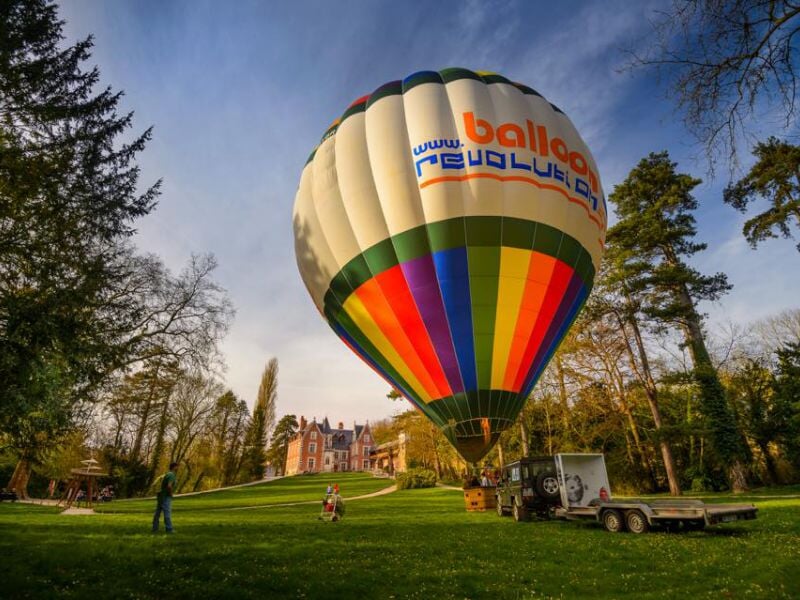 Montgolfière colorée s'élevant doucement au-dessus d'une pelouse verte, avec en arrière-plan un château entouré d'arbres, par une journée ensoleillée