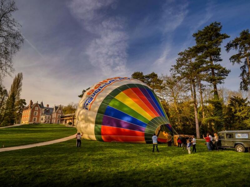 Montgolfière multicolore en cours de gonflage sur une pelouse verdoyante devant un château en briques, entourée de personnes observant la préparation sous un ciel bleu parsemé de nuages