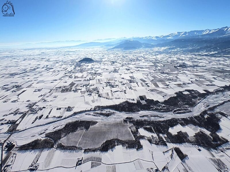 Vue aérienne d'un paysage hivernal avec champs enneigés et montagnes au loin sous un ciel bleu clair.