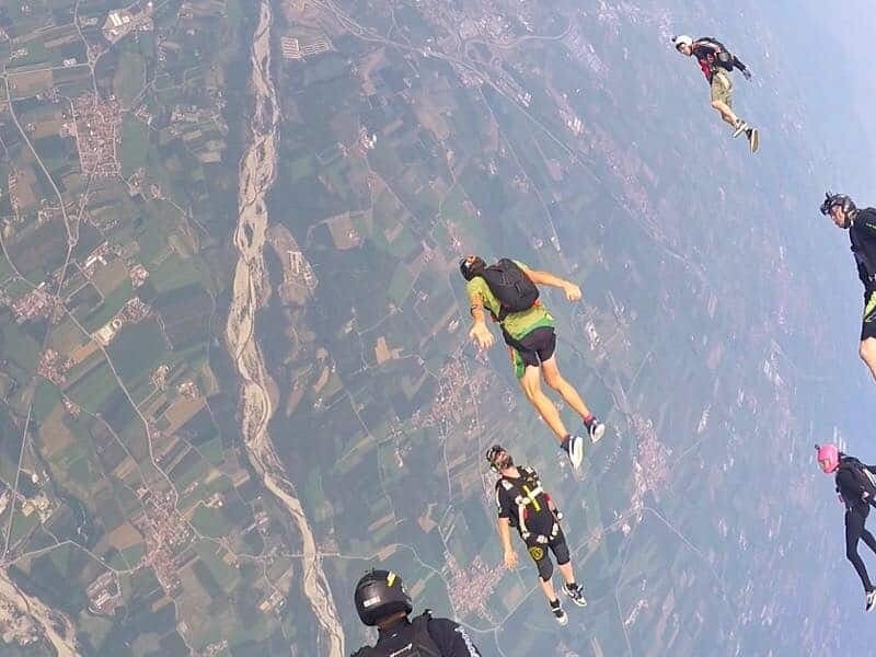 Plusieurs parachutistes en chute libre au-dessus d'un paysage rural avec champs et rivière visibles en contrebas.
