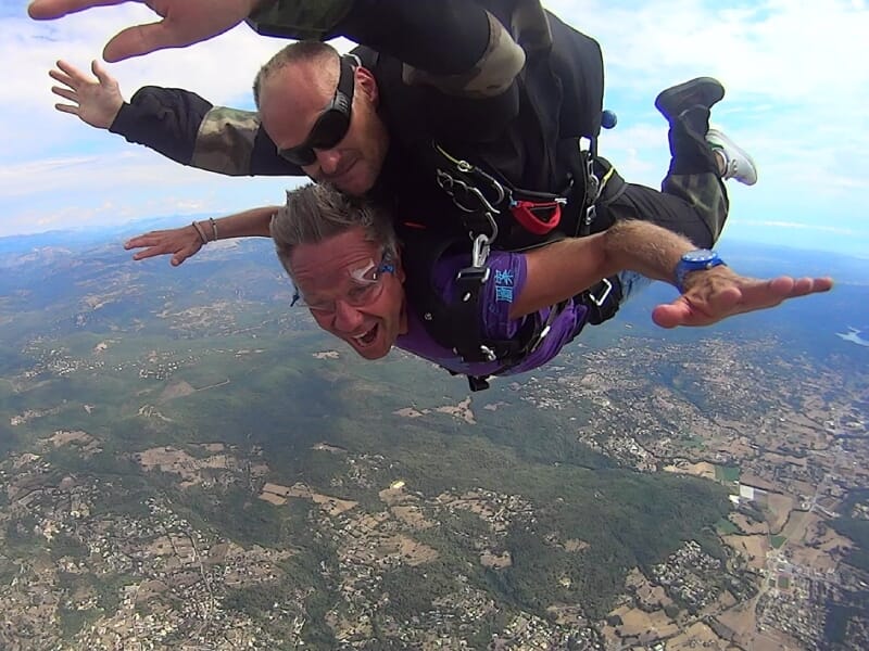 Saut en parachute en tandem, deux personnes en chute libre, sourire éclatant. Paysage rural avec champs, forêts et habitations en dessous. Ciel bleu avec quelques nuages. Activité extrême et adrénalin