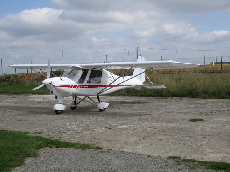 Petit avion ultraléger blanc stationné sur une piste en béton avec herbe et ciel nuageux en arrière-plan.
