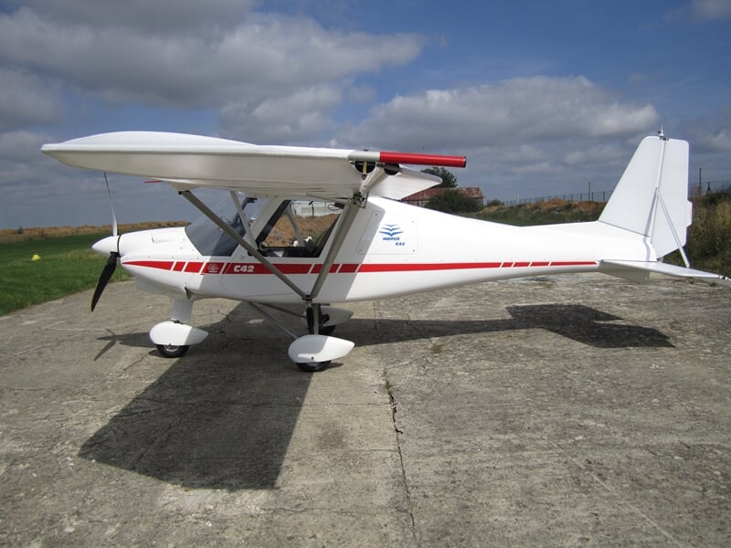 Avion ultraléger blanc avec une bande rouge garé sur une piste en béton sous un ciel partiellement nuageux.