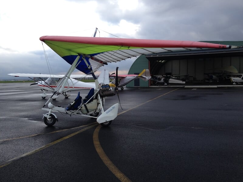 ULM biplace avec ailes colorées rouge, vert et bleu sur une piste humide devant un hangar vert.